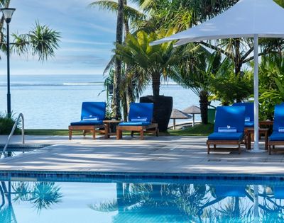 Poolside scene with four blue loungers under a large white umbrella, overlooking a tranquil ocean. Tropical palm trees create a serene, relaxing ambiance.