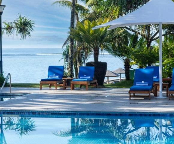 Poolside scene with four blue loungers under a large white umbrella, overlooking a tranquil ocean. Tropical palm trees create a serene, relaxing ambiance.