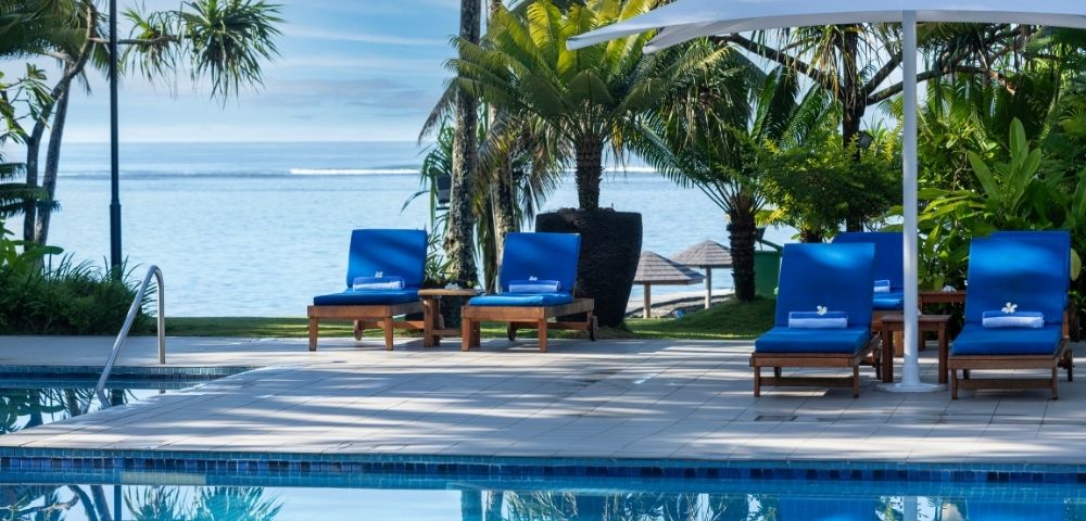 Poolside scene with four blue loungers under a large white umbrella, overlooking a tranquil ocean. Tropical palm trees create a serene, relaxing ambiance.