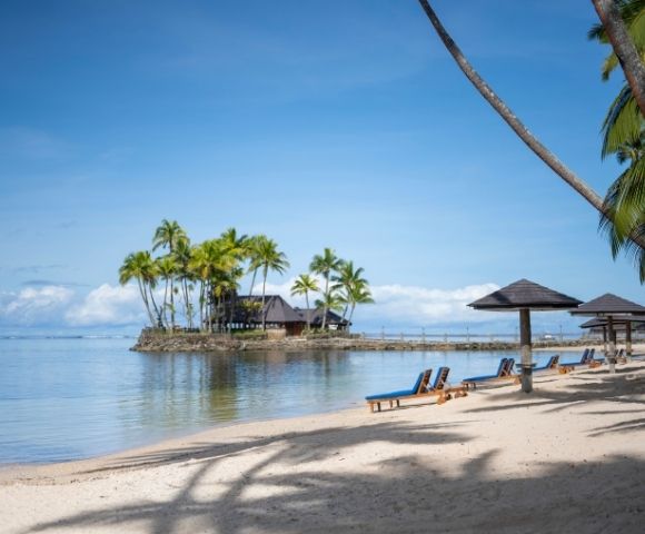 Tropical beach scene with empty lounge chairs under wooden canopies on sandy shore. Palm trees and clear blue sky create a serene and relaxing atmosphere.