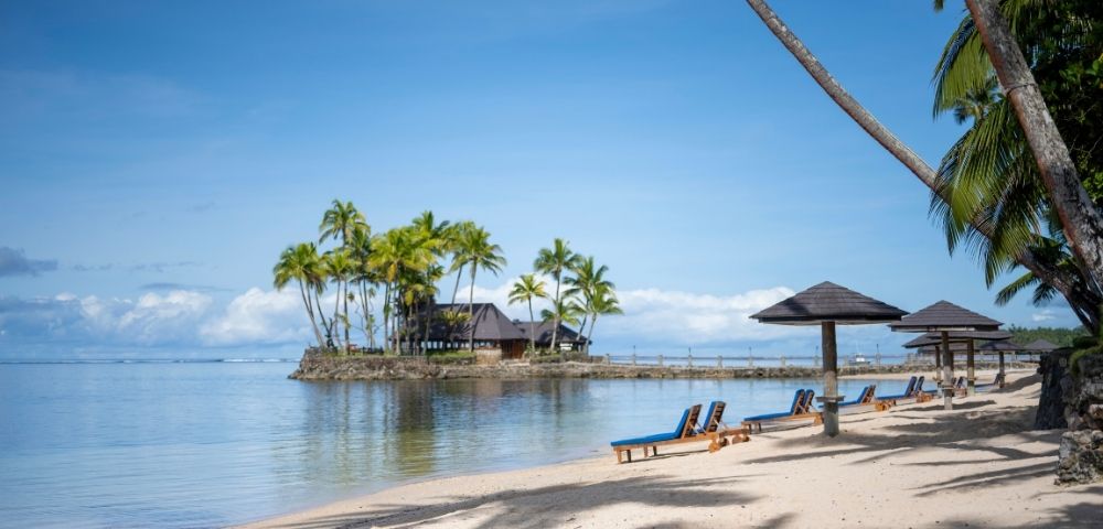 Tropical beach scene with empty lounge chairs under wooden canopies on sandy shore. Palm trees and clear blue sky create a serene and relaxing atmosphere.