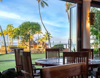 Open-air restaurant with wooden tables and chairs overlooking a tropical beach view. Palm trees and a thatched gazebo by the water under a bright sky.
