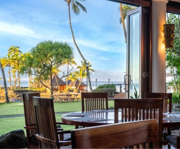 Open-air restaurant with wooden tables and chairs overlooking a tropical beach view. Palm trees and a thatched gazebo by the water under a bright sky.v