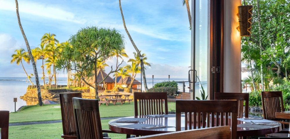 Open-air restaurant with wooden tables and chairs overlooking a tropical beach view. Palm trees and a thatched gazebo by the water under a bright sky.