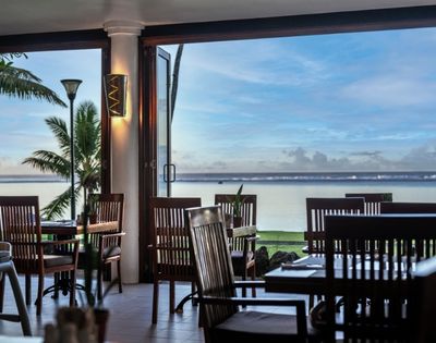 Open-air restaurant with wooden chairs and tables, overlooking a serene ocean view at sunset. Palm trees frame the scene, creating a tranquil, relaxing ambiance.