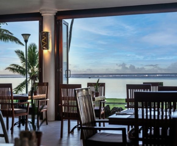 Open-air restaurant with wooden chairs and tables, overlooking a serene ocean view at sunset. Palm trees frame the scene, creating a tranquil, relaxing ambiance.