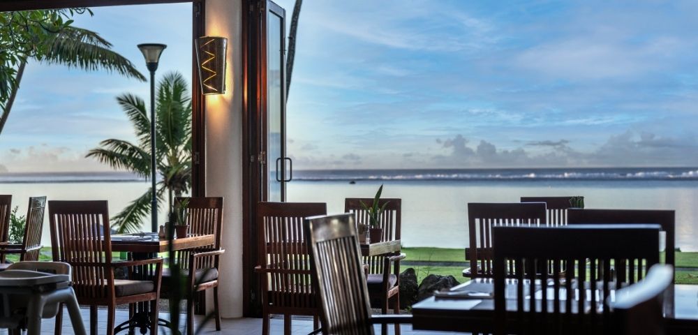 Open-air restaurant with wooden chairs and tables, overlooking a serene ocean view at sunset. Palm trees frame the scene, creating a tranquil, relaxing ambiance.