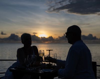A couple clinks wine glasses at a table overlooking the ocean during a vibrant sunset. Silhouetted figures add a romantic and peaceful tone.