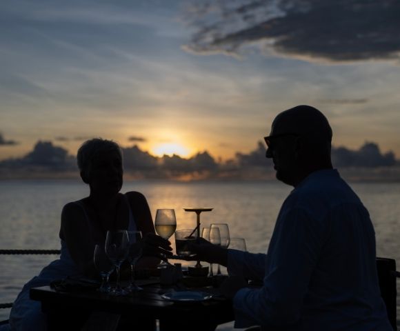 A couple clinks wine glasses at a table overlooking the ocean during a vibrant sunset. Silhouetted figures add a romantic and peaceful tone.