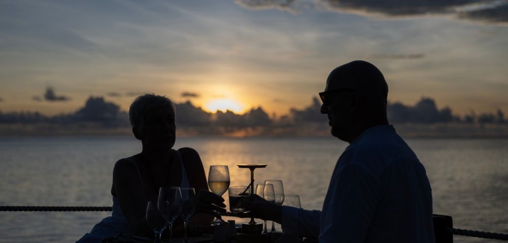A couple clinks wine glasses at a table overlooking the ocean during a vibrant sunset. Silhouetted figures add a romantic and peaceful tone.