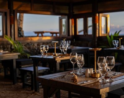 Cozy beachfront restaurant at sunset, featuring wooden tables set with elegant glassware and lamps. Warm lighting and open windows reveal ocean views.