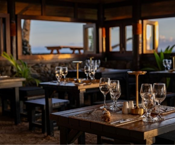 Cozy beachfront restaurant at sunset, featuring wooden tables set with elegant glassware and lamps. Warm lighting and open windows reveal ocean views.