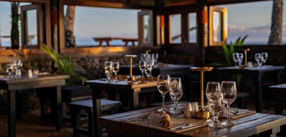 Cozy beachfront restaurant at sunset, featuring wooden tables set with elegant glassware and lamps. Warm lighting and open windows reveal ocean views.