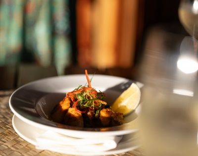 A gourmet dish on a white plate, garnished with herbs and a lemon wedge, set on a woven placemat. A glass of white wine is slightly blurred in the foreground.