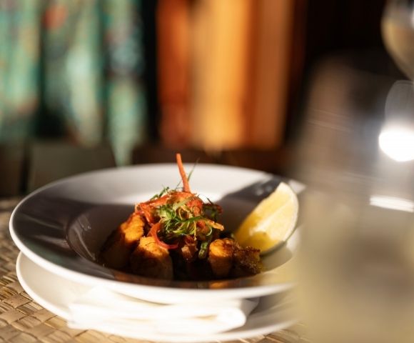 A gourmet dish on a white plate, garnished with herbs and a lemon wedge, set on a woven placemat. A glass of white wine is slightly blurred in the foreground.
