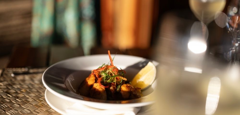 A gourmet dish on a white plate, garnished with herbs and a lemon wedge, set on a woven placemat. A glass of white wine is slightly blurred in the foreground.