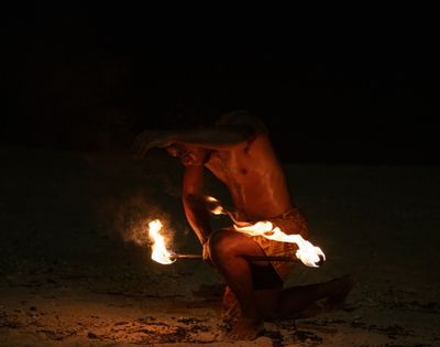 A shirtless performer, kneeling on sand, maneuvers flaming torches in a captivating fire dance at night, exuding intensity and concentration in the dark.