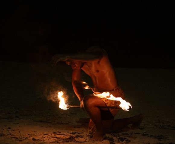 A shirtless performer, kneeling on sand, maneuvers flaming torches in a captivating fire dance at night, exuding intensity and concentration in the dark.