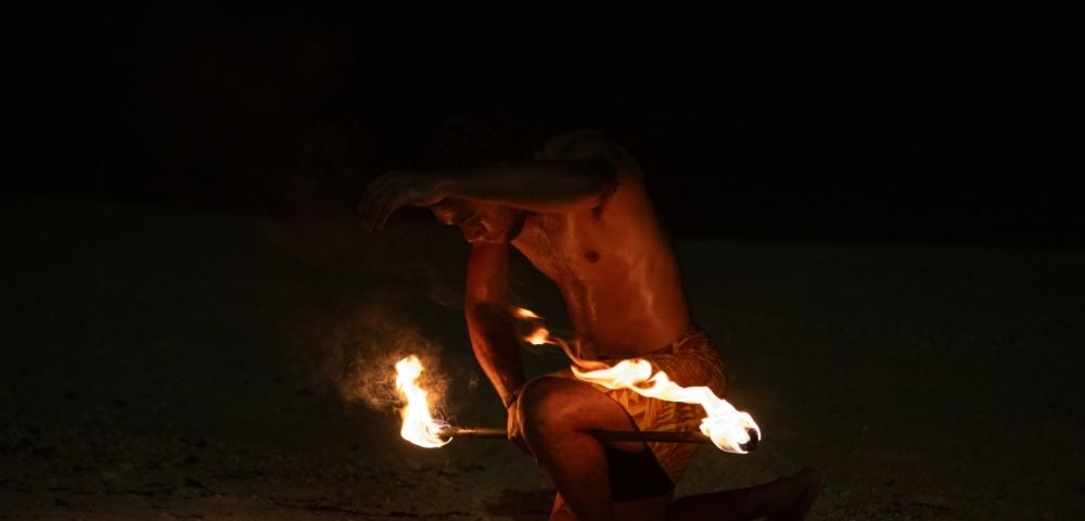 A shirtless performer, kneeling on sand, maneuvers flaming torches in a captivating fire dance at night, exuding intensity and concentration in the dark.