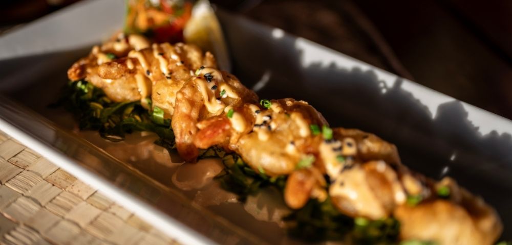 A close-up of a gourmet dish featuring grilled seafood topped with sauce and sesame seeds, on a bed of greens. Bright, appetizing presentation.