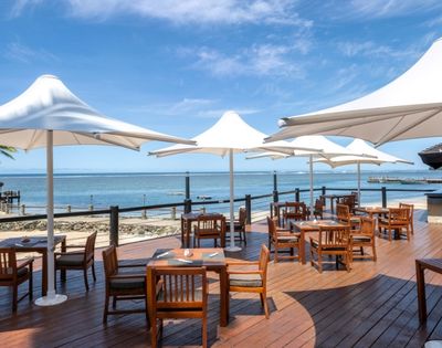 Outdoor dining area with wooden tables and chairs under large white umbrellas on a sunny beachside deck. The calm sea and clear blue sky are in the background.