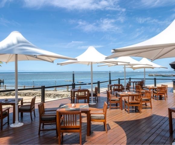 Outdoor dining area with wooden tables and chairs under large white umbrellas on a sunny beachside deck. The calm sea and clear blue sky are in the background.