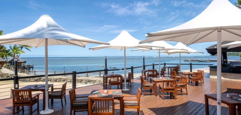 Outdoor dining area with wooden tables and chairs under large white umbrellas on a sunny beachside deck. The calm sea and clear blue sky are in the background.