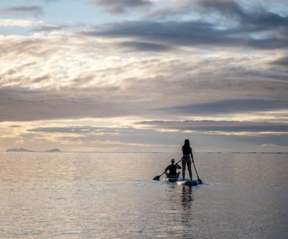 Two people paddleboarding on calm ocean waters during a serene sunset, with a cloudy sky and distant islands on the horizon, creating a peaceful atmosphere.