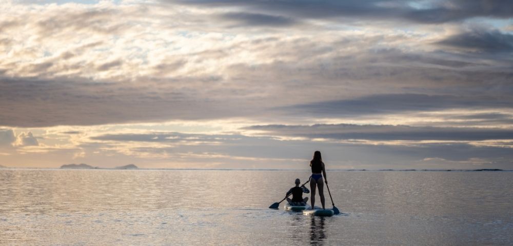 Two people paddleboarding on calm ocean waters during a serene sunset, with a cloudy sky and distant islands on the horizon, creating a peaceful atmosphere.