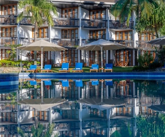 Luxurious hotel with white facade and numerous balconies, surrounded by lush greenery. Blue sun loungers sit by a tranquil pool, evoking relaxation.
