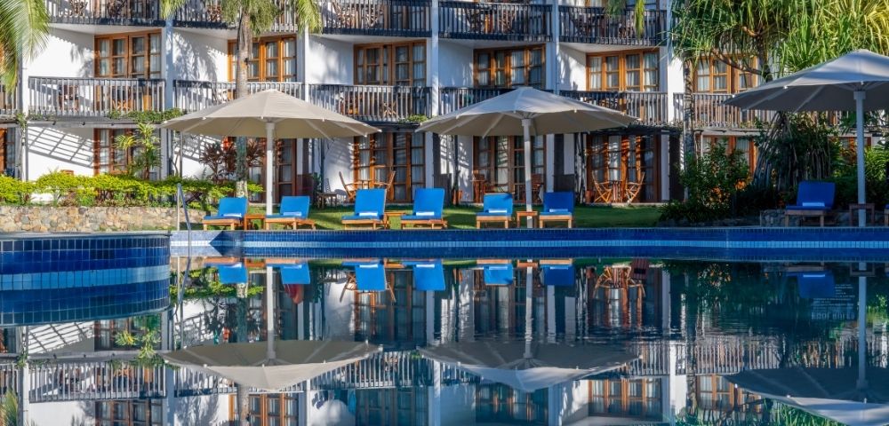 Luxurious hotel with white facade and numerous balconies, surrounded by lush greenery. Blue sun loungers sit by a tranquil pool, evoking relaxation.