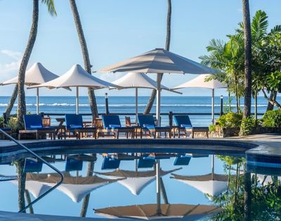 A serene tropical poolside scene with blue lounge chairs and white umbrellas reflecting in the water. Tall palm trees and the ocean are in the background.