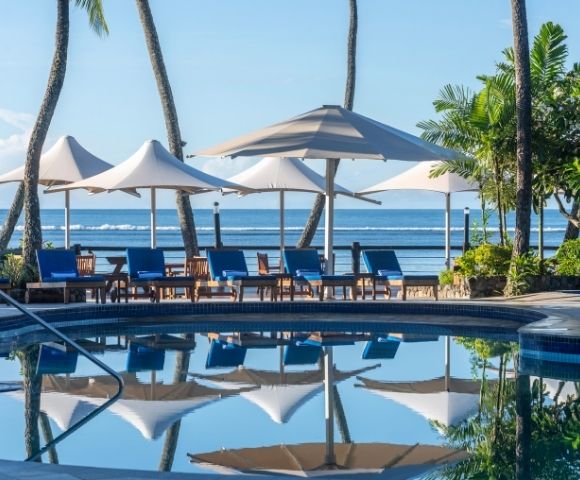 A serene tropical poolside scene with blue lounge chairs and white umbrellas reflecting in the water. Tall palm trees and the ocean are in the background.