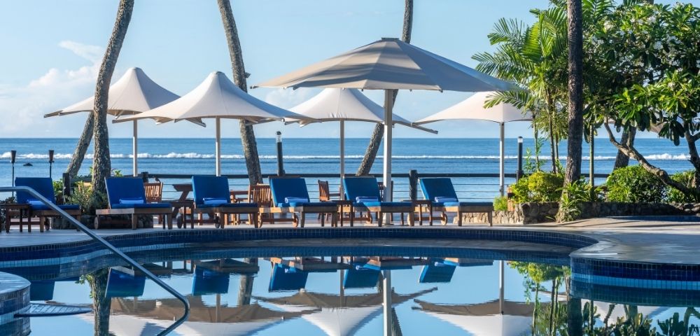 A serene tropical poolside scene with blue lounge chairs and white umbrellas reflecting in the water. Tall palm trees and the ocean are in the background.