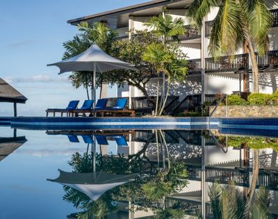 A serene poolside scene with a row of blue lounge chairs under a white umbrella, surrounded by lush trees. The calm water reflects the tropical setting.