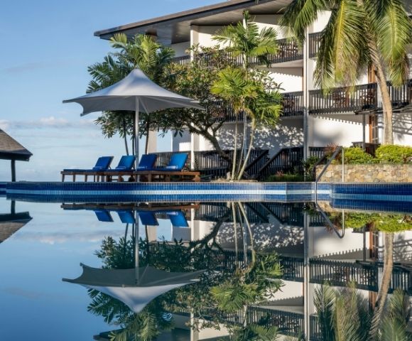 A serene poolside scene with a row of blue lounge chairs under a white umbrella, surrounded by lush trees. The calm water reflects the tropical setting.
