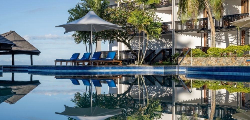 A serene poolside scene with a row of blue lounge chairs under a white umbrella, surrounded by lush trees. The calm water reflects the tropical setting.