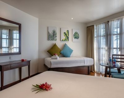 Bright, inviting hotel room with a modern decor. Features a double bed, single bed with green and blue pillows, leaf art, and a balcony. Relaxed ambiance.