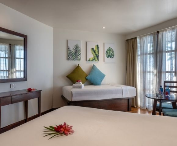 Bright, inviting hotel room with a modern decor. Features a double bed, single bed with green and blue pillows, leaf art, and a balcony. Relaxed ambiance.