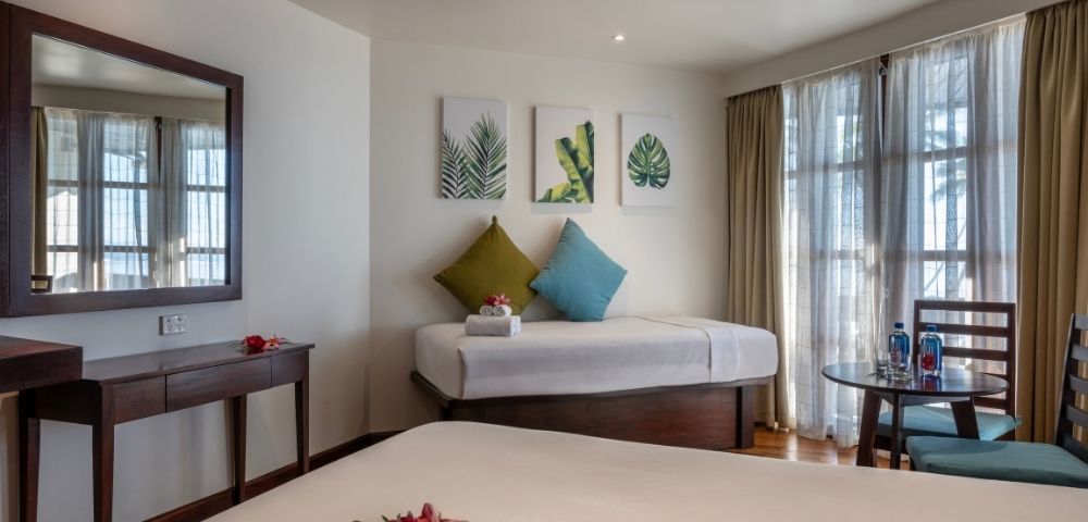 Bright, inviting hotel room with a modern decor. Features a double bed, single bed with green and blue pillows, leaf art, and a balcony. Relaxed ambiance.