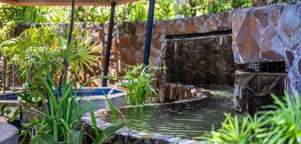 A serene garden scene with a stone wall waterfall flowing into a tranquil pond surrounded by lush green plants. A canopy provides shade, evoking peace.