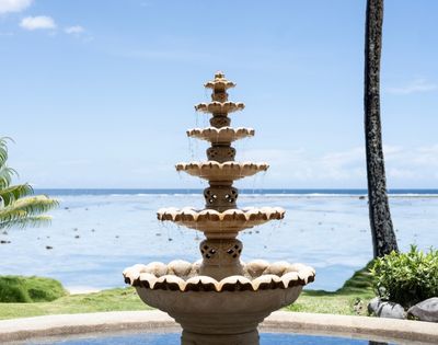 A tiered stone fountain with water cascading down sits by the ocean under a clear blue sky. Lush greenery surrounds the tranquil seaside scene.