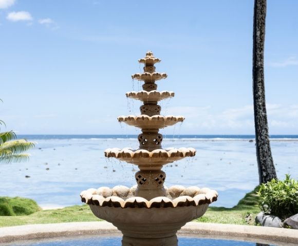 A tiered stone fountain with water cascading down sits by the ocean under a clear blue sky. Lush greenery surrounds the tranquil seaside scene.