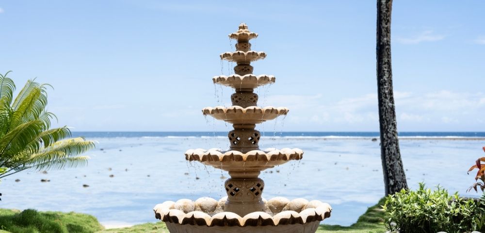 A tiered stone fountain with water cascading down sits by the ocean under a clear blue sky. Lush greenery surrounds the tranquil seaside scene.
