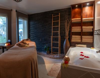 Cozy spa room featuring a massage table with brown towels, a stone wall, a ladder shelf with folded towels, and a bubble bath adorned with red petals.