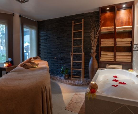 Cozy spa room featuring a massage table with brown towels, a stone wall, a ladder shelf with folded towels, and a bubble bath adorned with red petals.