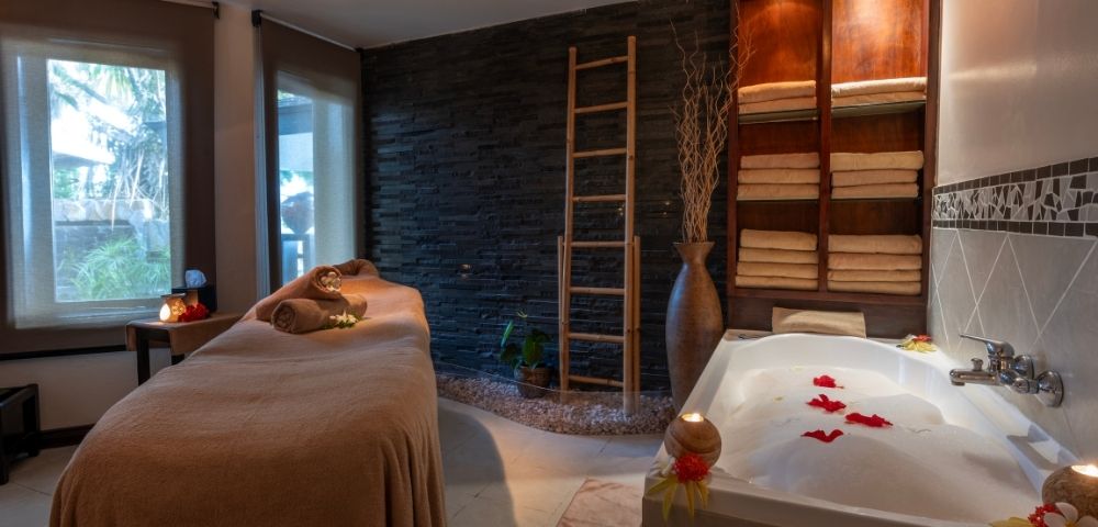 Cozy spa room featuring a massage table with brown towels, a stone wall, a ladder shelf with folded towels, and a bubble bath adorned with red petals.