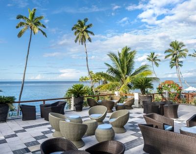 Scenic oceanfront patio with wicker chairs, tables, and lush palm trees under a blue sky. Calm ocean view creates a serene and tropical atmosphere.