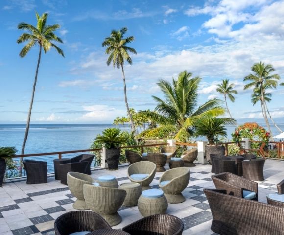 Scenic oceanfront patio with wicker chairs, tables, and lush palm trees under a blue sky. Calm ocean view creates a serene and tropical atmosphere.