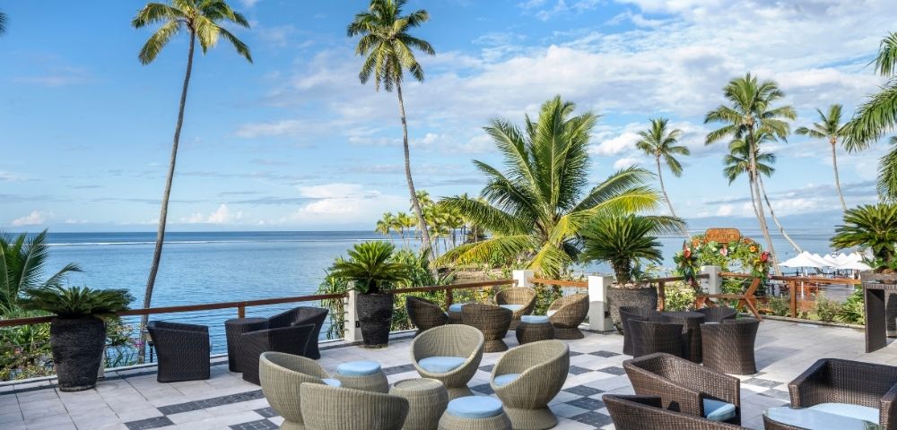 Scenic oceanfront patio with wicker chairs, tables, and lush palm trees under a blue sky. Calm ocean view creates a serene and tropical atmosphere.